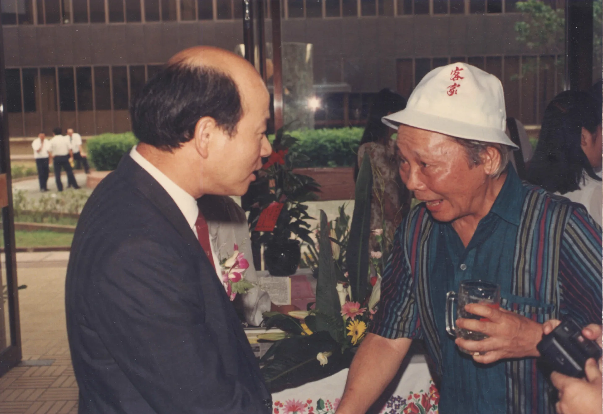 竹東陳天鵬醫師等人創辦新竹風雜誌創刊酒會，許信良（左）與鍾肇政（右）相見歡，鍾肇政戴的帽子第二屆客家夏令營學員們每人都有一頂。1991，新竹市，楊國鑫攝